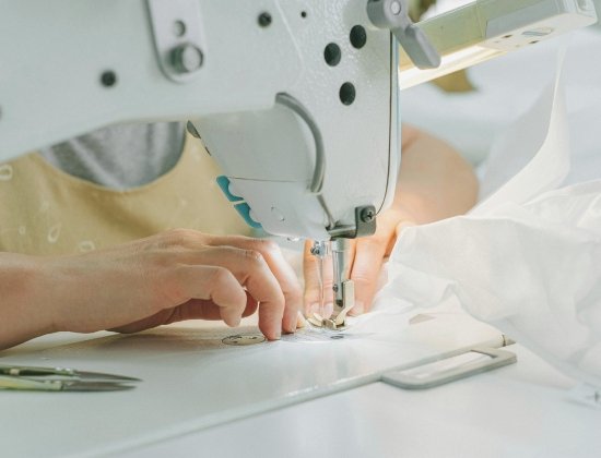 A close-up of a sewing machine in use, with two hands guiding white fabric under the needle; a pair of scissors sits on the sewing table.