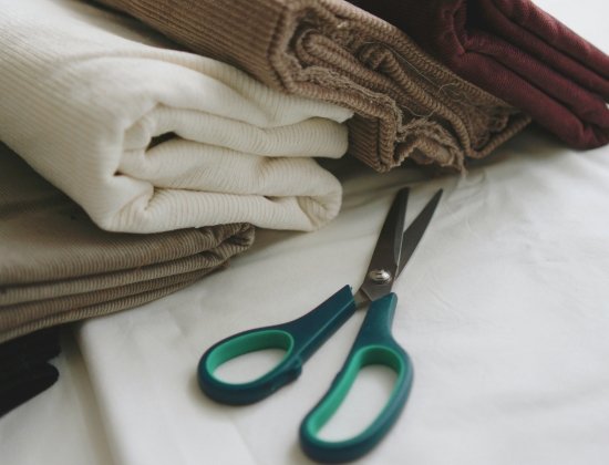 A close-up view of folded corduroy fabrics in beige, cream, brown, and burgundy, with a pair of green-handled scissors resting on a white surface.