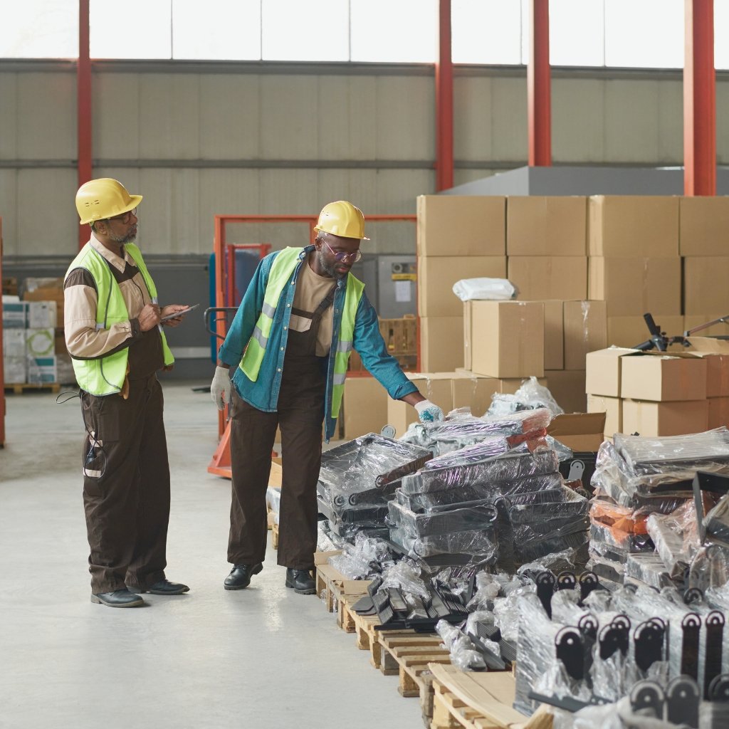 Two workers in yellow hard hats and safety vests inspect a pallet of wrapped parts in a warehouse, with cardboard boxes stacked in the background.